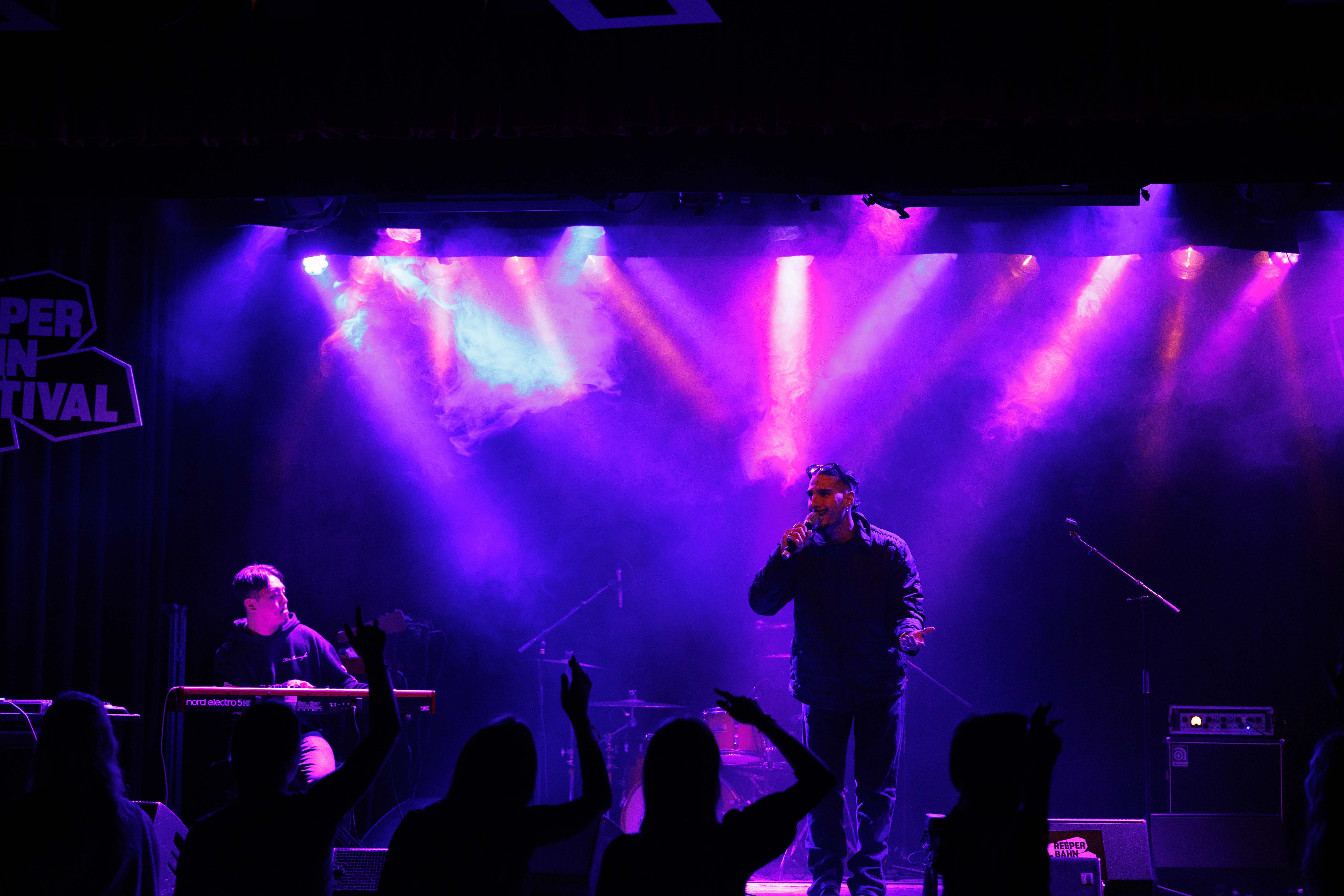 Ein Sänger auf einer Bühne mit lila Licht und Nebel, Publikum im Vordergrund.