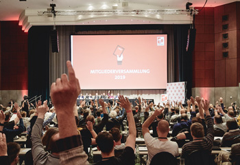 Teilnehmer einer Versammlung heben die Hände vor einer Bühne mit rotem Bildschirm und der Aufschrift