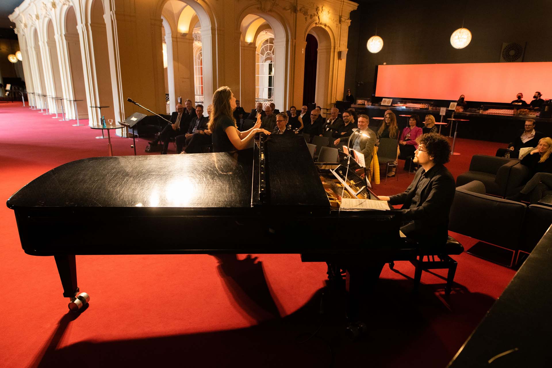Eine Frau singt neben einem Flügel, Pianist spielt, Publikum sitzt in einem eleganten Raum mit rotem Teppich.