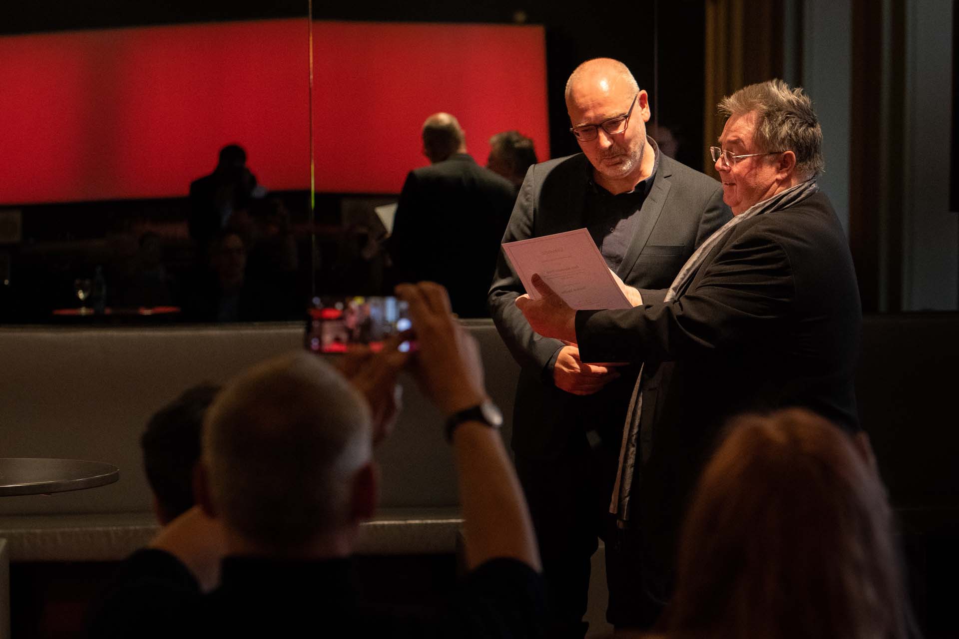 Zwei Männer in Anzügen betrachten ein Dokument in einem gedämpft beleuchteten Raum.