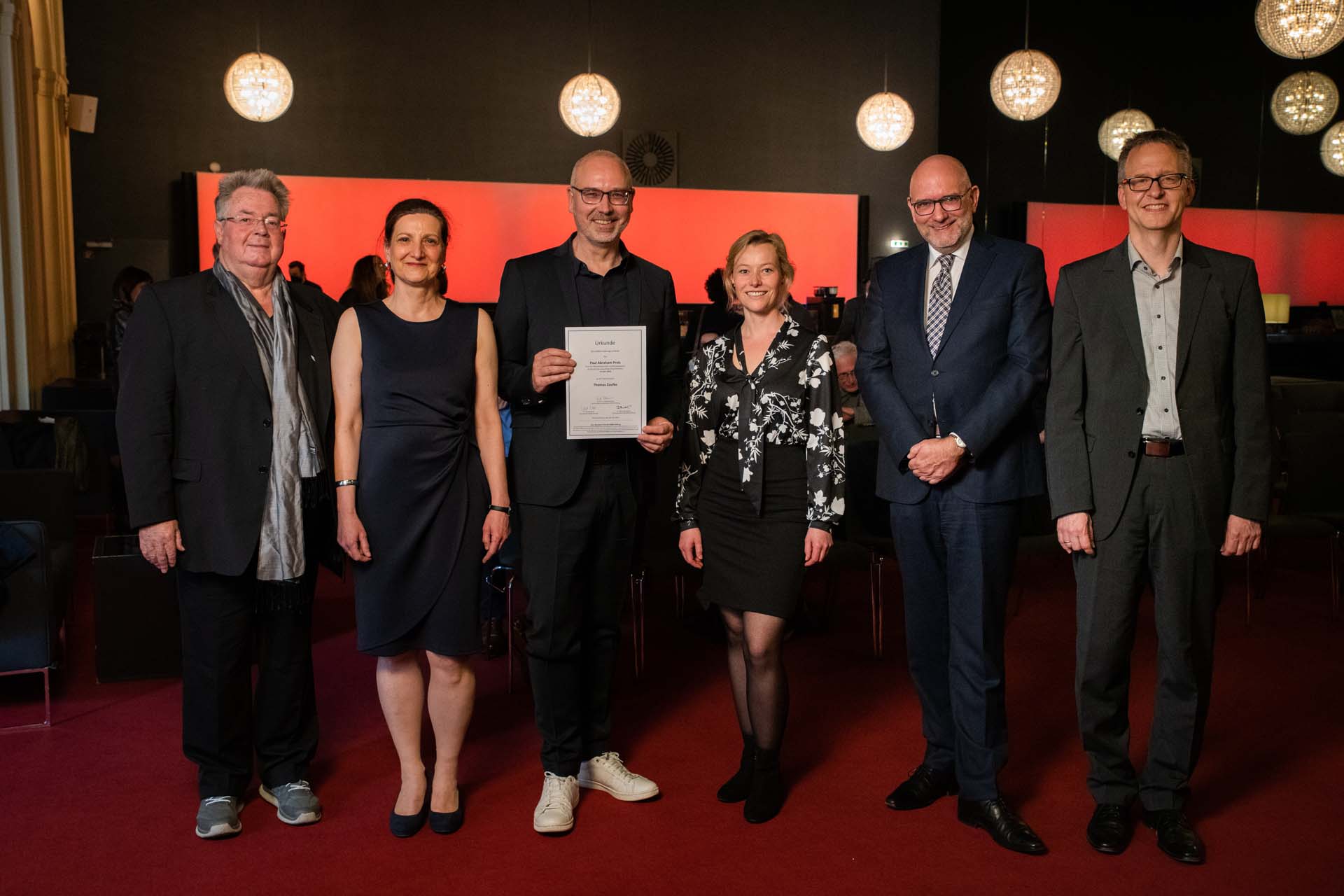 Sechs Personen in Abendkleidung stehen auf rotem Teppich, eine hält eine Urkunde. Hintergrund mit runden Leuchten.
