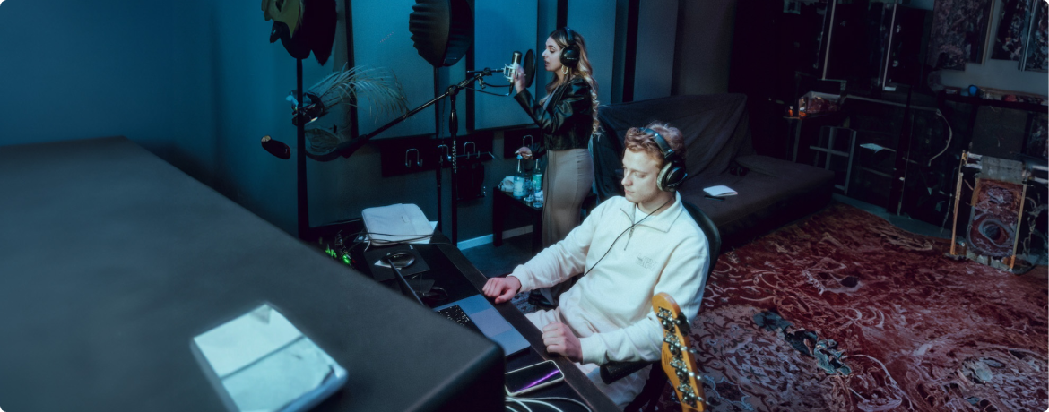 Eine Frau singt ins Mikrofon, während ein Mann am Laptop mit Kopfhörern im Tonstudio arbeitet.