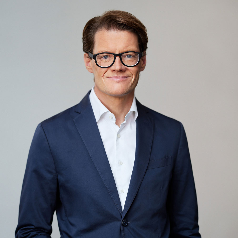 Ein Mann in Anzug und Brille lächelt vor neutralem Hintergrund.