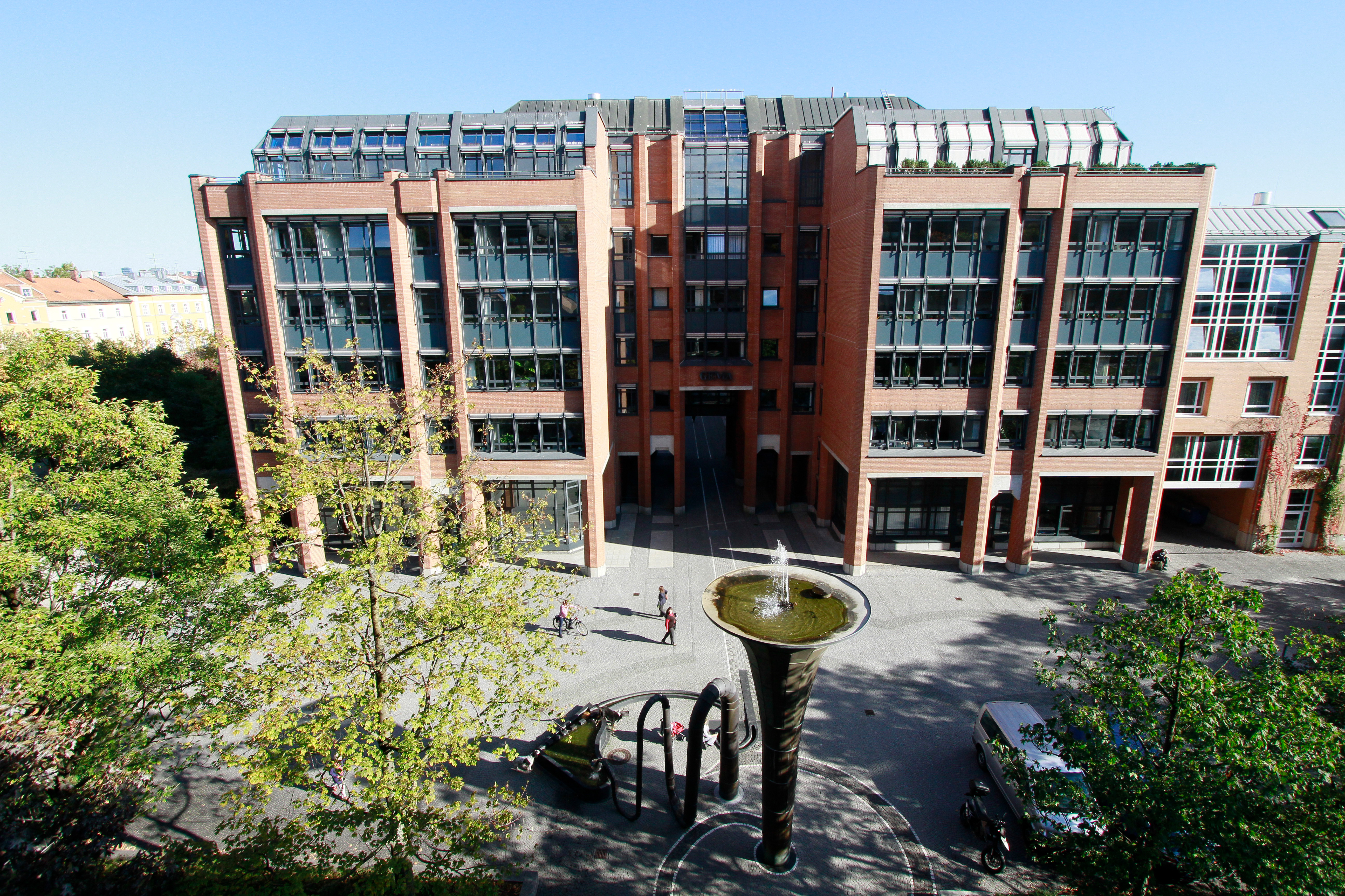Großes Backsteingebäude mit Glasfronten, davor ein Brunnen und Bäume, Menschen spazieren auf dem Platz.