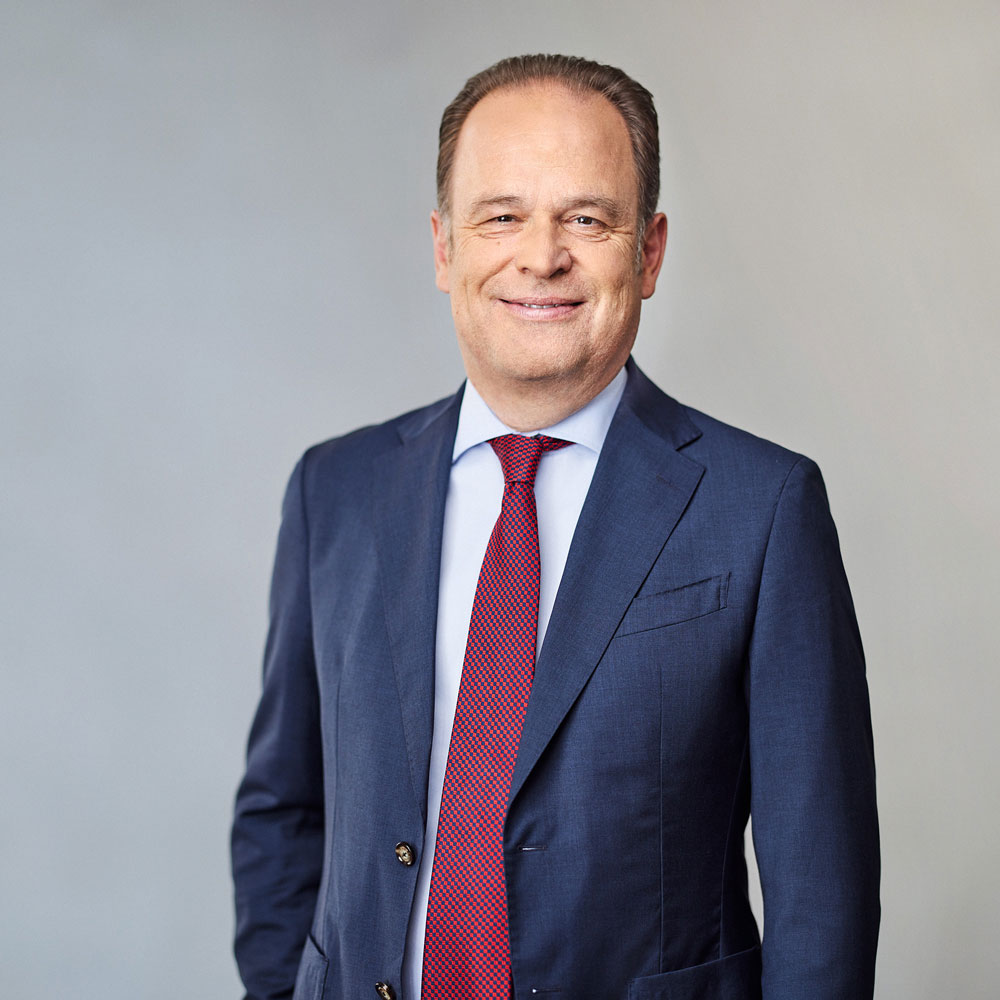 Ein Mann in blauem Anzug und roter Krawatte lächelt vor neutralem Hintergrund.