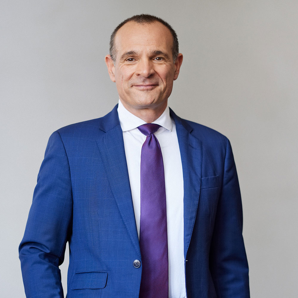 Ein Mann im blauen Anzug mit lila Krawatte lächelt vor einem neutralen Hintergrund.