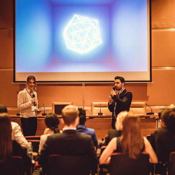 Zwei Personen präsentieren vor Publikum, hinter ihnen eine Leinwand mit geometrischem Muster.