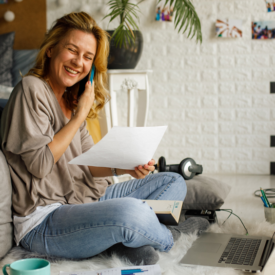 Eine Frau telefoniert lächelnd, sitzt auf dem Boden mit Laptop, Papieren und Hanteln umgeben.