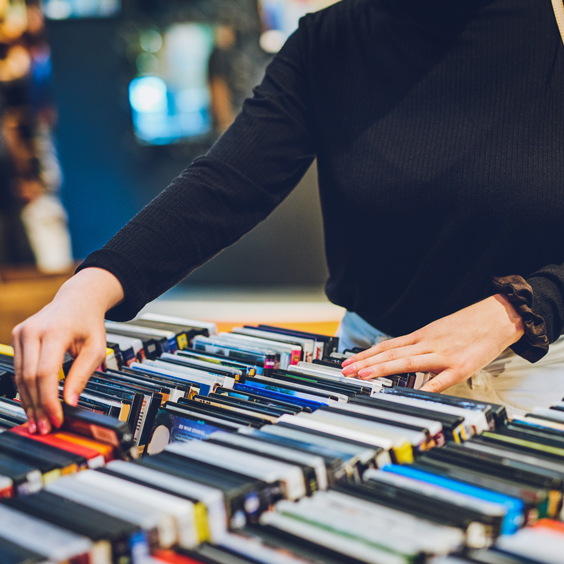 Person in schwarzem Pullover blättert durch gestapelte DVDs auf einem Tisch.