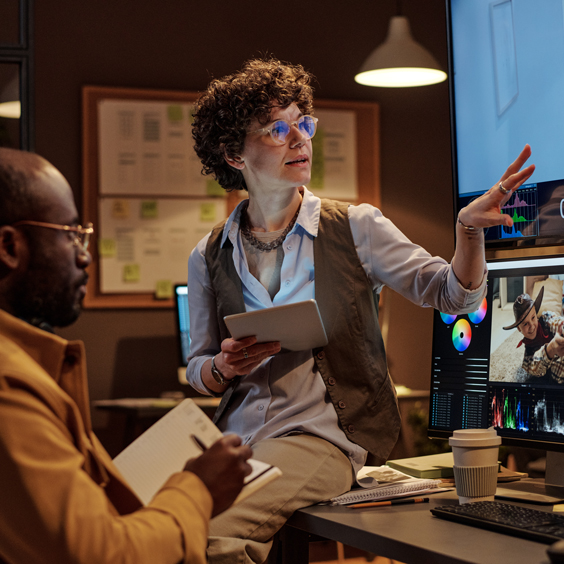 Eine Person zeigt auf einen Monitor, während eine andere Notizen macht