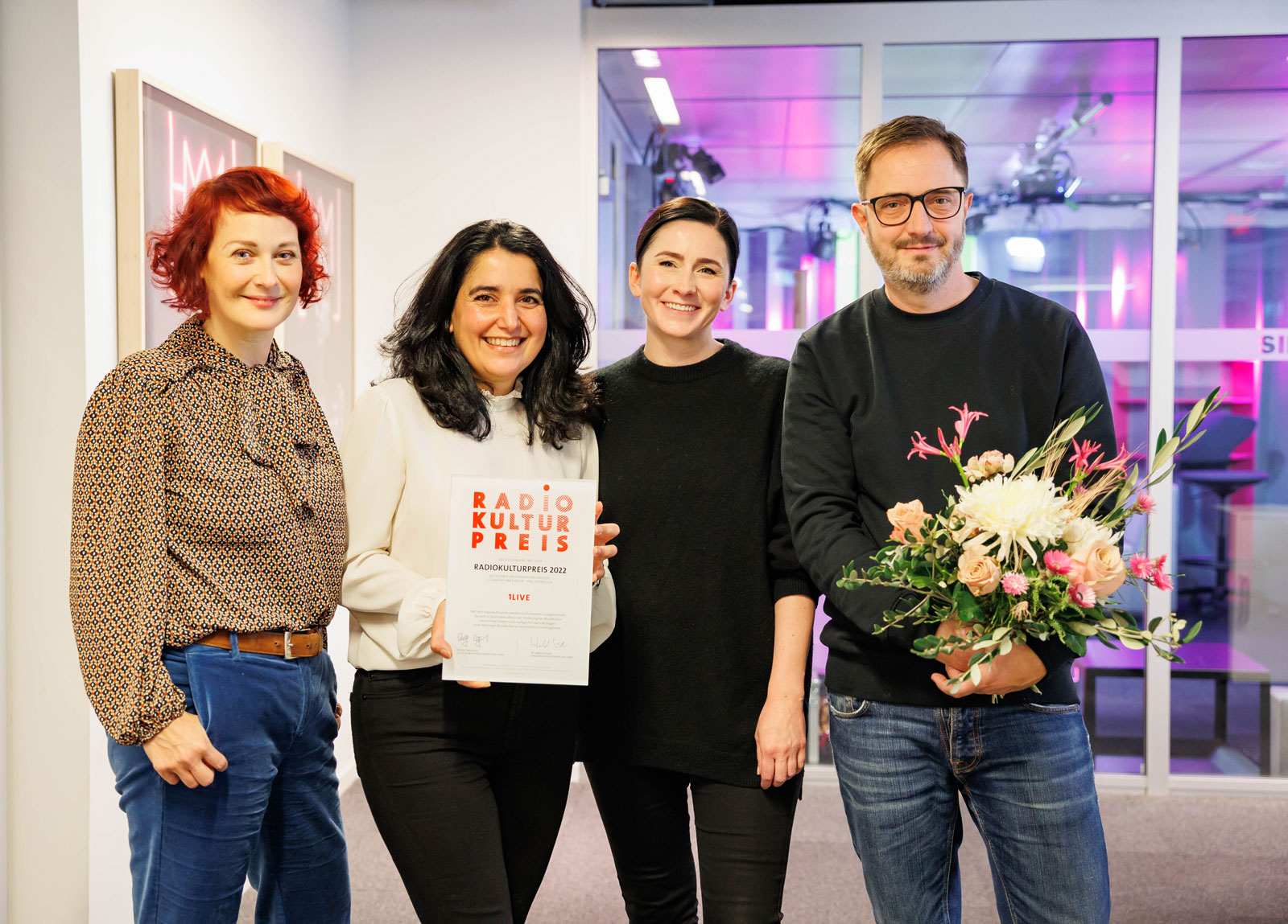 Vier Personen stehen lächelnd in einem Raum, eine hält eine Urkunde, eine andere einen Blumenstrauß.
