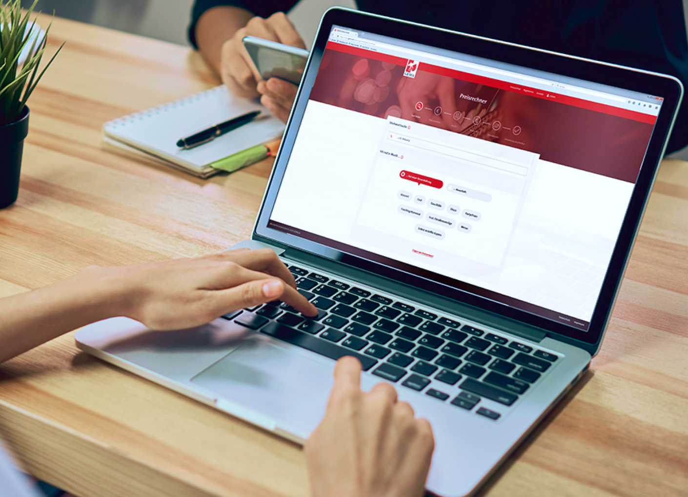 Eine Person tippt auf einem Laptop, daneben ein Notizbuch und ein Smartphone auf einem Holztisch.