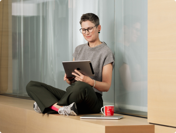 Eine Person sitzt auf einem Holzfensterbrett, liest ein Tablet, neben einer roten Tasse und einem Laptop.