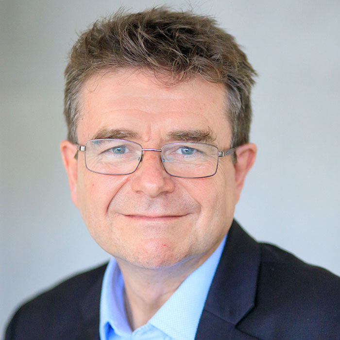 Ein lächelnder Mann mit Brille und Anzug vor grauem Hintergrund.