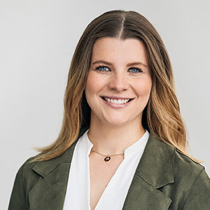 Eine lächelnde Frau mit langen Haaren, trägt eine grüne Jacke und eine weiße Bluse vor neutralem Hintergrund.