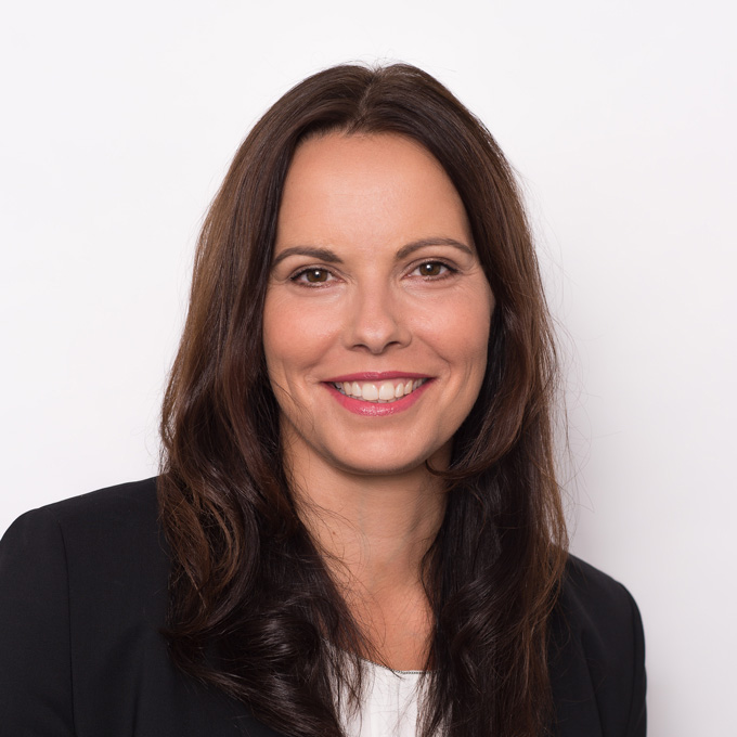 Eine lächelnde Frau mit langen braunen Haaren vor weißem Hintergrund. Sie trägt ein schwarzes Jackett.